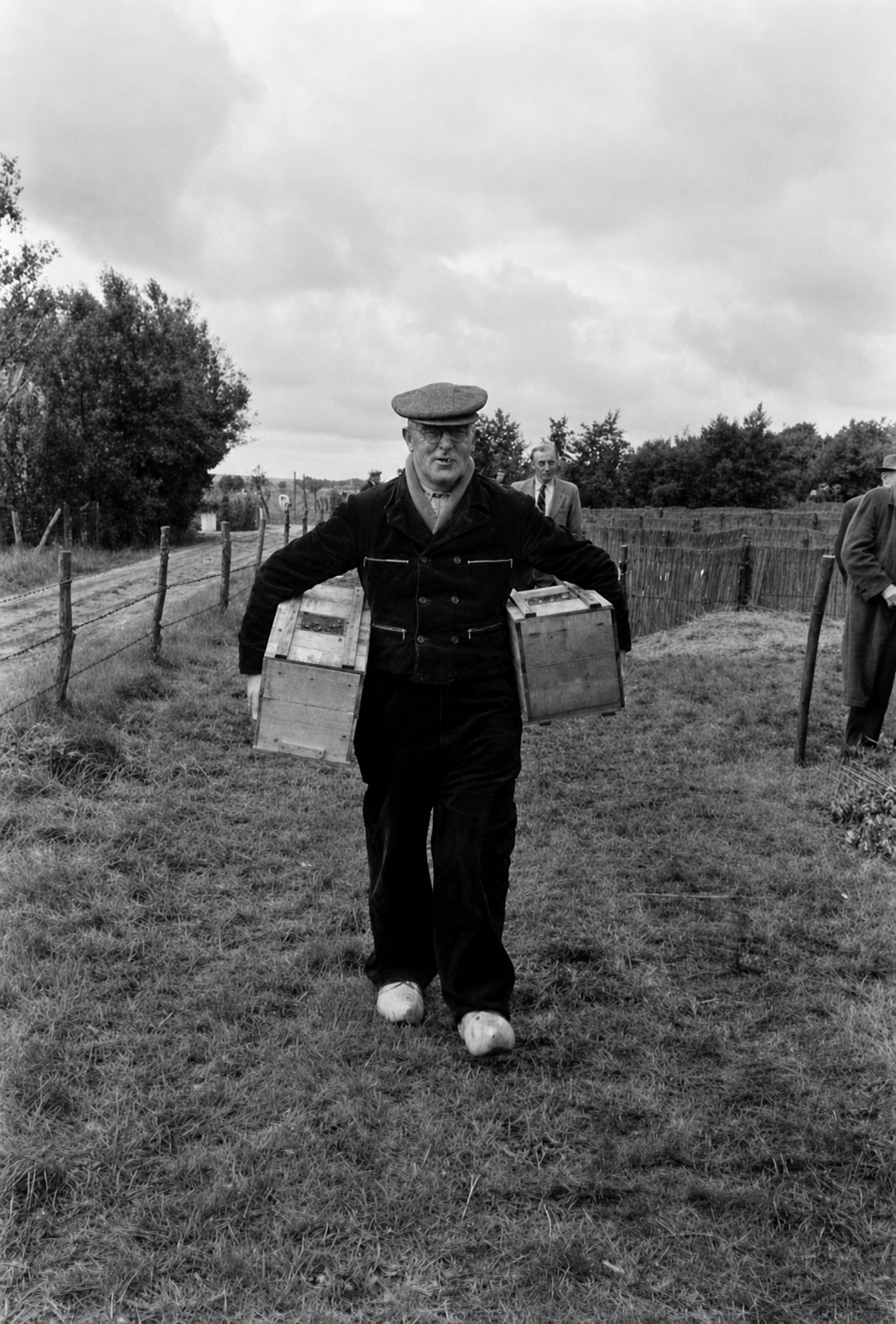 A beekeeper walking towards the camera with a wooden hive box under each arm