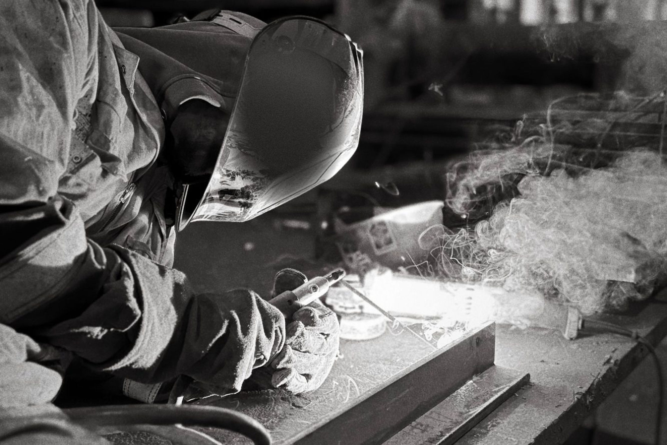 A welder bent over a steel bench, sparks and smoke lifting into the dark of the shop